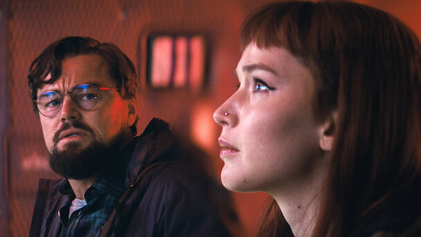 Two individuals in contemplative poses with a dimly lit, red-tinged interior in the background. The man has a beard and glasses, wearing a dark jacket, and the woman has long straight hair, staring intently ahead.